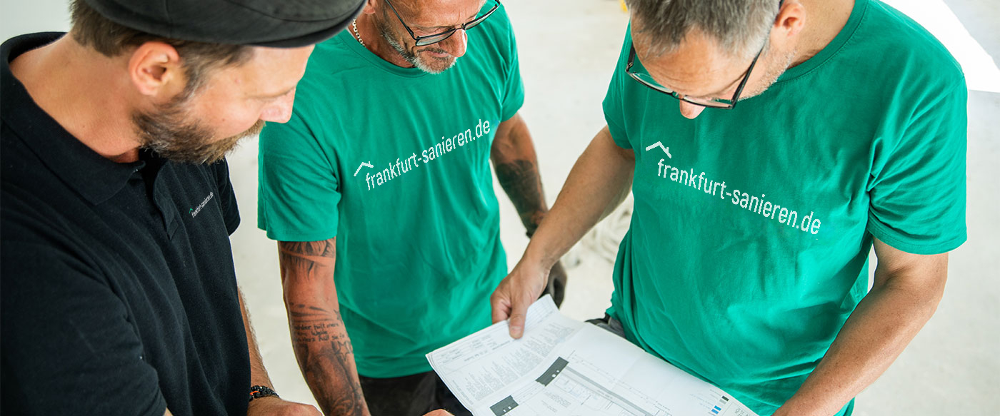 Drei Bauprofis von frankfurt-sanieren.de besprechen Baupläne auf einer Baustelle, mit Fokus auf professionelle Sanierungsplanung.