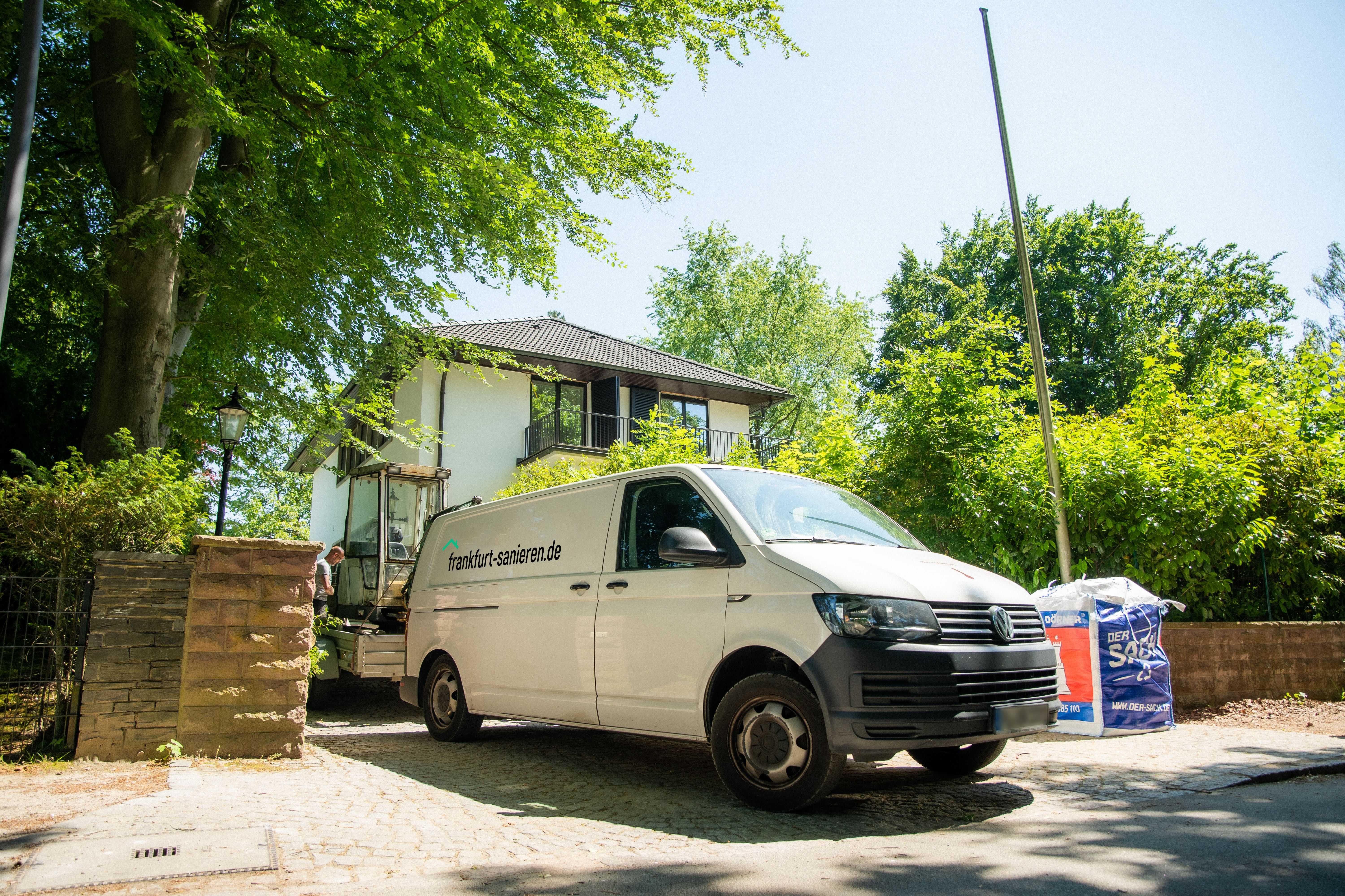 Transporter von frankfurt-sanieren.de vor einem Haus, bereit für Sanierungsarbeiten, umgeben von Bäumen und Baustellenmaterial.