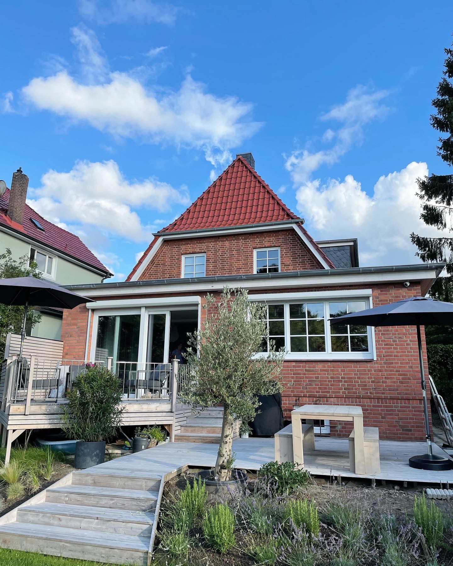 Modernisiertes rotes Backsteinhaus mit rotem Dach, großer Terrasse aus Holz, Gartenpflanzen, Sitzbereich, Sonnenschirmen und Treppenstufen unter blauem Himmel.